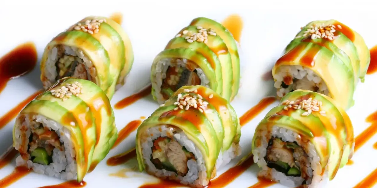 Sześć kawałków sushi dragon roll, udekorowanych plastrami awokado, sezamem i sosem.