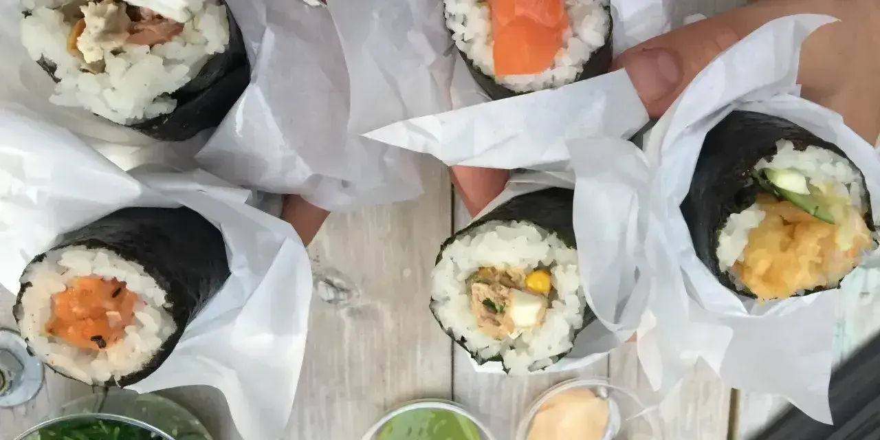 Ręce trzymają cztery pyszne temaki zawinięte w papier. Widać różne nadzienia: łososia, awokado, warzywa i rybę.