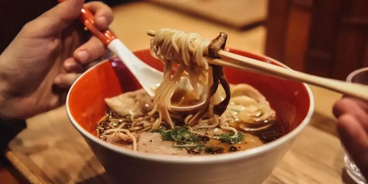 Ręka trzyma pałeczkami porcję parującego ramenu z makaronem i grzybami. Oto jak jesc ramen!