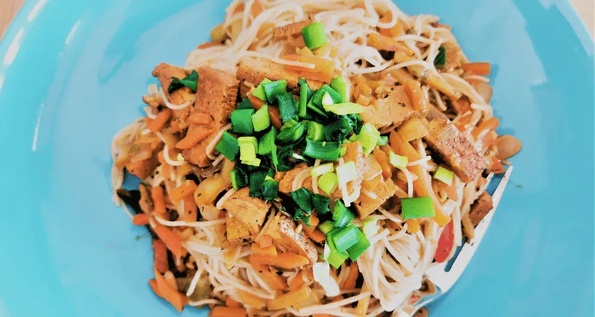 Pyszne tofu po chińsku z makaronem ryżowym, warzywami i szczypiorkiem na niebieskim talerzu.