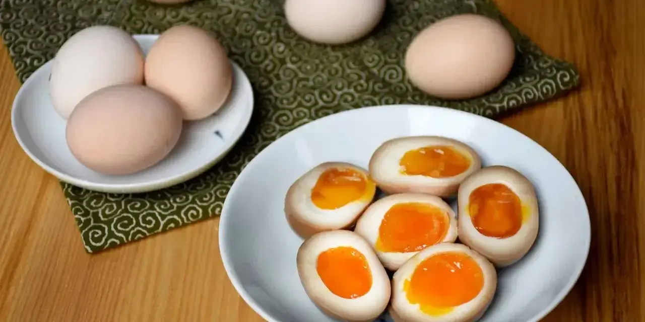 Na talerzu połówki jajek z płynnym, pomarańczowym żółtkiem, idealne jajka do ramenu. Obok całe jajka.