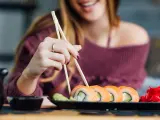 Uśmiechnięta kobieta w ciąży sięga po sushi. Na talerzu rolki z łososiem i awokado, obok wasabi i marynowany imbir.