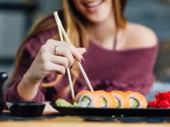 Uśmiechnięta kobieta w ciąży sięga po sushi. Na talerzu rolki z łososiem i awokado, obok wasabi i marynowany imbir.