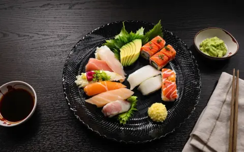 Talerz z różnorodnym sushi: nigiri z łososiem, tuńczykiem, kalmarem, rolki z łososiem, awokado. Obok sos sojowy, wasabi i pałeczki. Czy sushi się podgrzewa? Nie, to danie serwuje się na zimno.