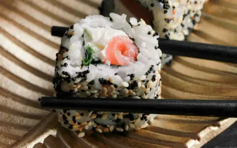 Rolka sushi z łososiem i serkiem, posypana sezamem, trzymana pałeczkami. Czy sushi można jeść na drugi dzień? Tak, jeśli było odpowiednio przechowywane.