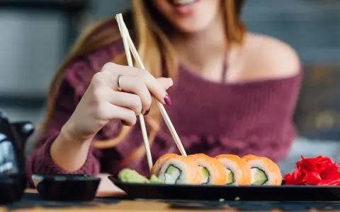 Uśmiechnięta kobieta w ciąży sięga po sushi. Na talerzu rolki z łososiem i awokado, obok wasabi i marynowany imbir.