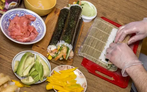 Przygotowanie sushi z warzywami: ryż, awokado, mango, krewetki i marynowany imbir. Ręce w rękawiczkach zwijają rolkę.