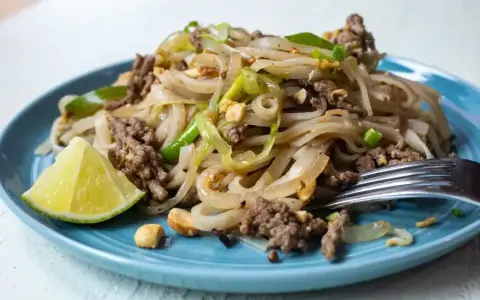 Pyszny pad thai z wołowiną, makaronem ryżowym, orzeszkami ziemnymi i ćwiartką limonki na niebieskim talerzu.