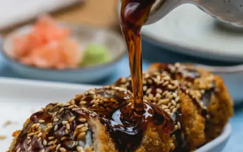Polewany sosem tajskim, chrupiący rolka sushi z sezamem. Obok talerzyk z imbirem i wasabi.