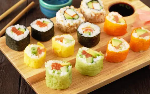 Kolorowe rolki sushi, w tym te z łososiem i awokado, gotowe do zawijania sushi, leżą na drewnianej desce.