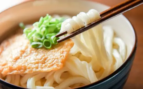 Miseczka z makaronem udon, polana sosem, posypana szczypiorkiem. Pałeczki unoszą porcję grubego makaronu. Pyszne danie, które warto znać pod kątem makaron udon kcal.
