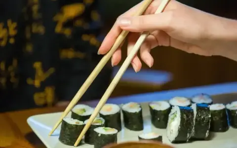 Dłoń podnosi pałeczkami rolkę sushi. Na talerzu ułożone są kawałki sushi, każdy z nich ma około 30 kcal.