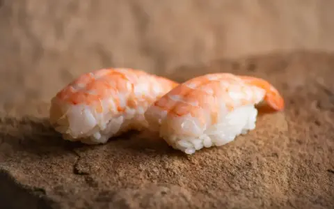 Dwa kawałki sushi z ryżem i różowymi krewetkami do sushi, ułożone na kamiennej powierzchni.