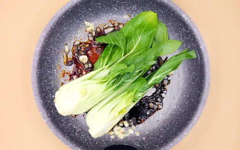 Na patelni smaży się aromatyczny sos z czosnkiem i kawałkami cebuli, a na nim leżą świeże liście sałaty pak choi.