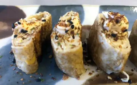 Trzy kawałki sushi z jajkiem, posypane czarnym sezamem i chrupiącymi dodatkami.