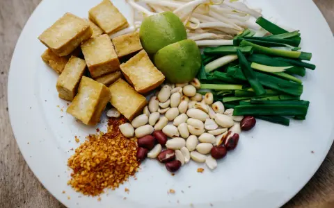 Na talerzu składniki do pad thai: smażone tofu, orzeszki ziemne, szczypiorek, kiełki fasoli mung, limonki i płatki chili. Gotowy sos pad thai czeka!