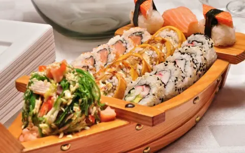 Drewniana łódka pełna sushi, przypominająca o tym, skąd pochodzi sushi. Obok stos białych talerzyków.