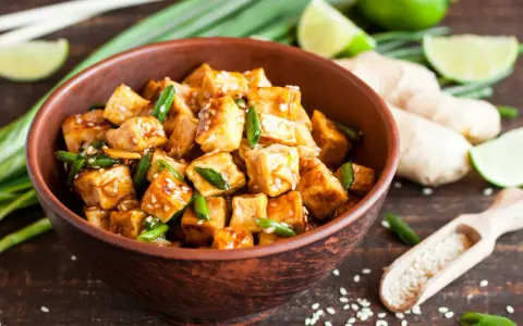 Pokrojone w kostkę, glazurowane tofu z zieloną cebulką i sezamem w misce. Obok limonka, imbir i drewniana łyżka z sezamem.