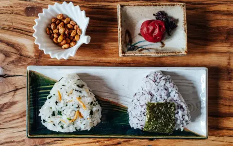 Dwa onigiri z różnym nadzieniem do onigiri: jedno z zielonymi dodatkami i rybkami, drugie z fioletowym ryżem i nori. Obok miseczka orzeszków i marynowana śliwka.