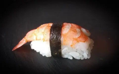 Nigiri ebi, czyli sushi z krewetką, na czarnym tle. Krewetka jest ułożona na ryżu, a całość spięta paskiem nori.