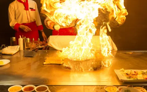Pokaz teppanyaki z płomieniami buchającymi z płyty grillowej. Kucharz w czerwonym fartuchu dodaje składniki, tworząc widowiskowy spektakl.