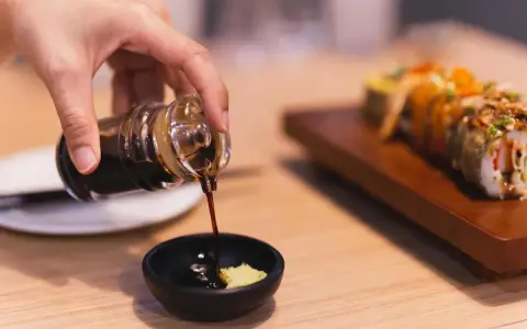 Dłoń nalewa sos sojowy bezglutenowy do miseczki z wasabi. Obok leży sushi.