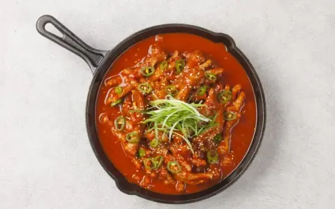 Kurczak w sosie gochujang, z zieloną cebulką i sezamem, podany na patelni.