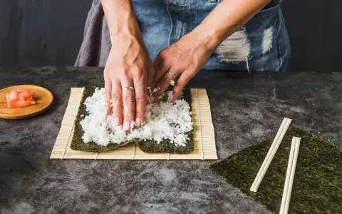 Ręce rozkładają ryż na nori, przygotowując się do zwinięcia sushi. Obok leżą pałeczki i talerzyk z imbirem, idealny dodatek do sushi.