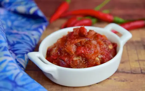 Pikantny sos sambal oelek w białej miseczce, obok czerwonych papryczek chili i niebieskiej tkaniny.
