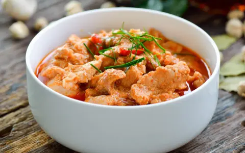 Aromaticzne curry z wieprzowiną w białej misce, ozdobione zieloną szczypiorkiem i czerwonymi papryczkami chili.
