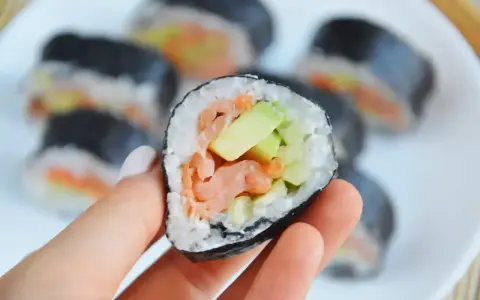 Hosomaki łosoś z awokado, trzymane w dłoni, na talerzu pełnym sushi.