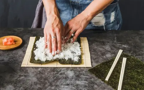 Dłonie rozprowadzają ryż na nori. Obok leżą pałeczki i marynowany imbir. To klucz do idealnego ryżu do sushi, który potrzebuje odrobiny octu.