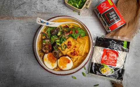 Szybki ramen przepis: miska pełna aromatycznego bulionu z grzybami shiitake, tofu, jajkiem i szczypiorkiem. Obok opakowanie makaronu i grzybów.
