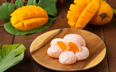 Słodkie mochi o smaku mango, ułożone na drewnianym talerzu, obok świeżych owoców i liści.
