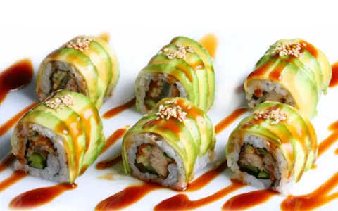 Sześć kawałków sushi dragon roll, udekorowanych plastrami awokado, sezamem i sosem.