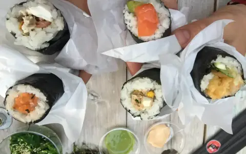 Ręce trzymają cztery pyszne temaki zawinięte w papier. Widać różne nadzienia: łososia, awokado, warzywa i rybę.