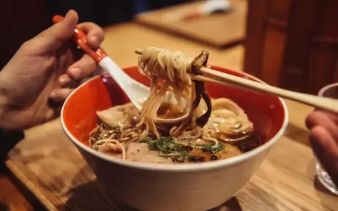 Ręka trzyma pałeczkami porcję parującego ramenu z makaronem i grzybami. Oto jak jesc ramen!
