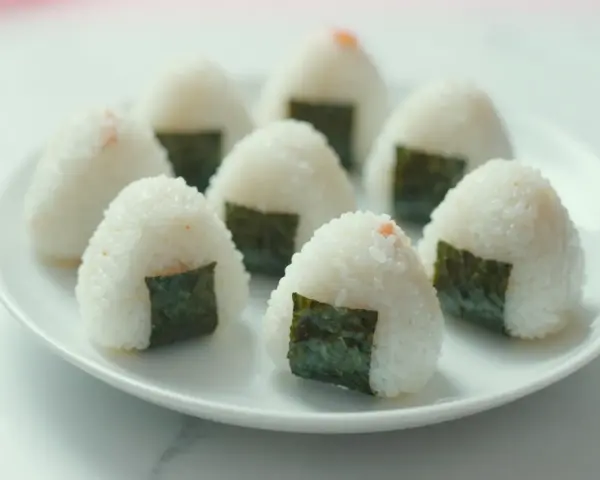 Kilka pysznych onigiri z łososiem, udekorowanych kawałkami nori, ułożonych na białym talerzu.