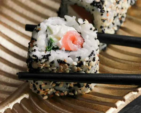 Rolka sushi z łososiem i serkiem, posypana sezamem, trzymana pałeczkami. Czy sushi można jeść na drugi dzień? Tak, jeśli było odpowiednio przechowywane.