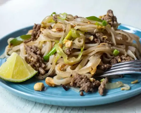 Pyszny pad thai z wołowiną, makaronem ryżowym, orzeszkami ziemnymi i ćwiartką limonki na niebieskim talerzu.