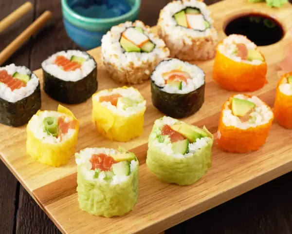 Kolorowe rolki sushi, w tym te z łososiem i awokado, gotowe do zawijania sushi, leżą na drewnianej desce.
