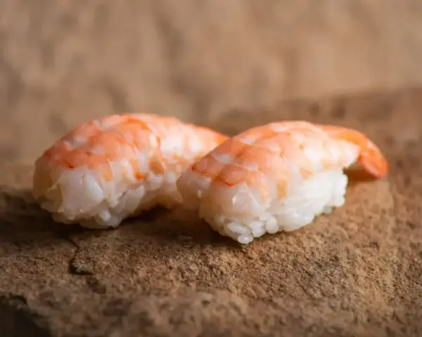 Dwa kawałki sushi z ryżem i różowymi krewetkami do sushi, ułożone na kamiennej powierzchni.