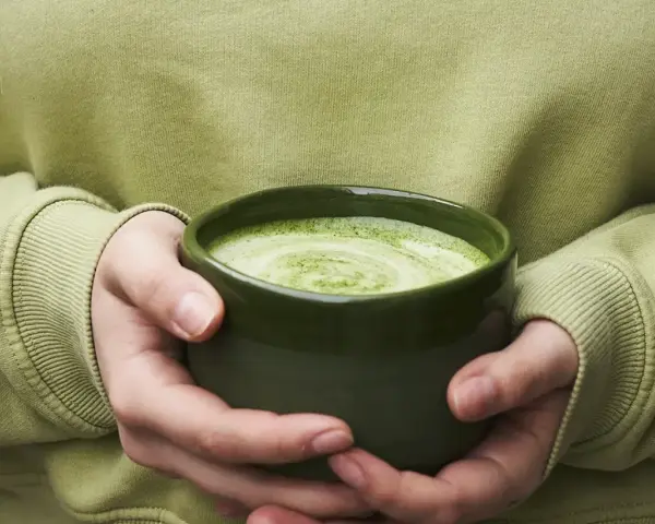 Dłonie trzymające zieloną miseczkę z pianką matcha.