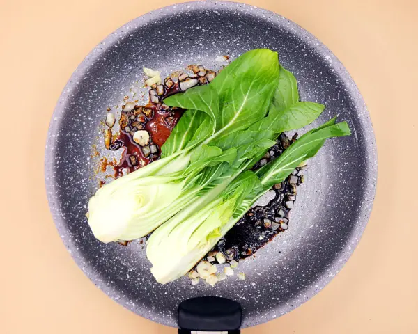 Na patelni smaży się aromatyczny sos z czosnkiem i kawałkami cebuli, a na nim leżą świeże liście sałaty pak choi.