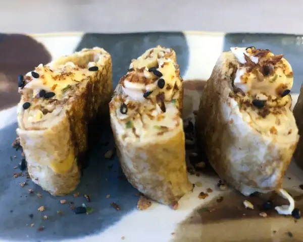 Trzy kawałki sushi z jajkiem, posypane czarnym sezamem i chrupiącymi dodatkami.