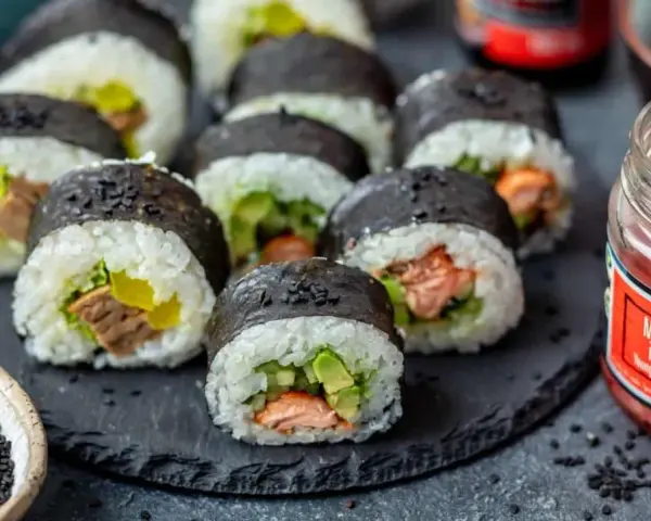 Pyszne sushi z łososiem, awokado i ogórkiem, posypane czarnym sezamem. Obok słoiczek marynowanego imbiru.