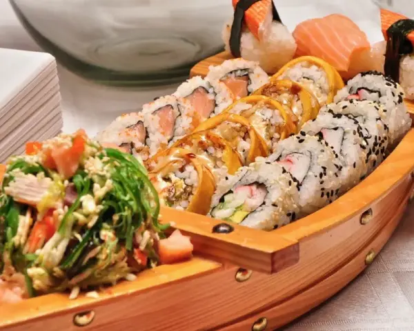Drewniana łódka pełna sushi, przypominająca o tym, skąd pochodzi sushi. Obok stos białych talerzyków.