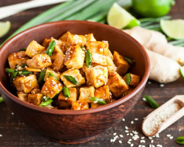 Pokrojone w kostkę, glazurowane tofu z zieloną cebulką i sezamem w misce. Obok limonka, imbir i drewniana łyżka z sezamem.