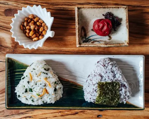 Dwa onigiri z różnym nadzieniem do onigiri: jedno z zielonymi dodatkami i rybkami, drugie z fioletowym ryżem i nori. Obok miseczka orzeszków i marynowana śliwka.