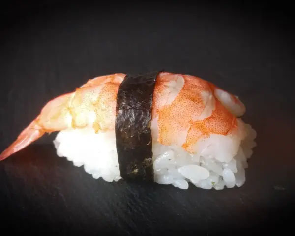 Nigiri ebi, czyli sushi z krewetką, na czarnym tle. Krewetka jest ułożona na ryżu, a całość spięta paskiem nori.
